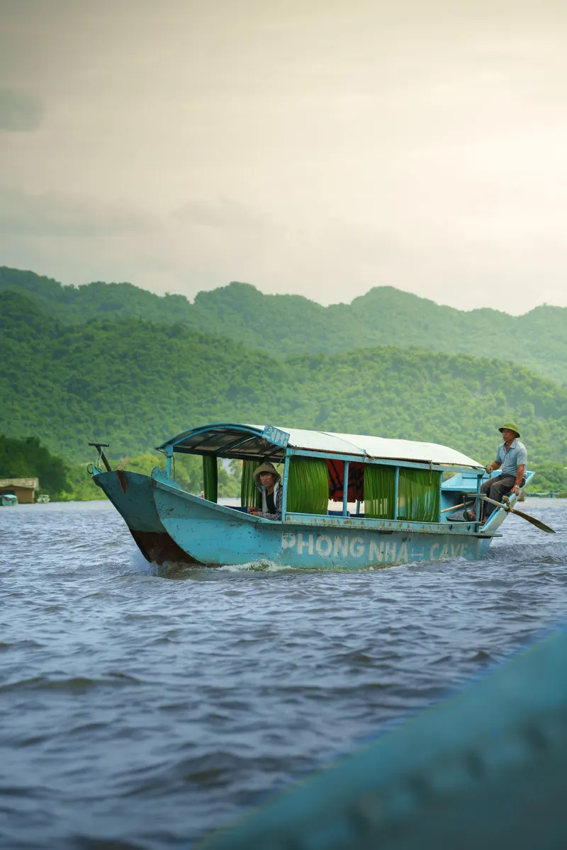 A boatman steers a blue Phong Nha Cave tour boat with a passenger along the Son River, green hills behind