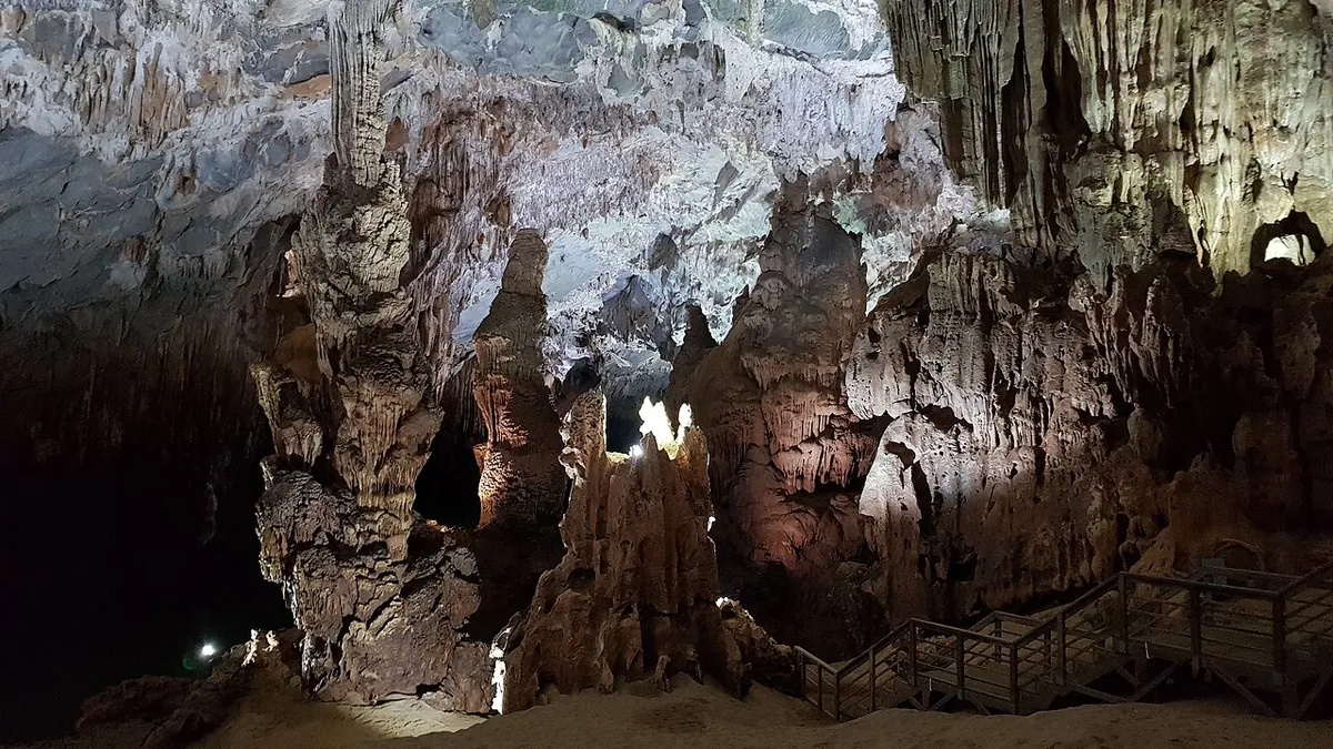Lit stalactites and stalagmites in a wide dry chamber of Phong Nha Cave, with a boardwalk railing along the floor