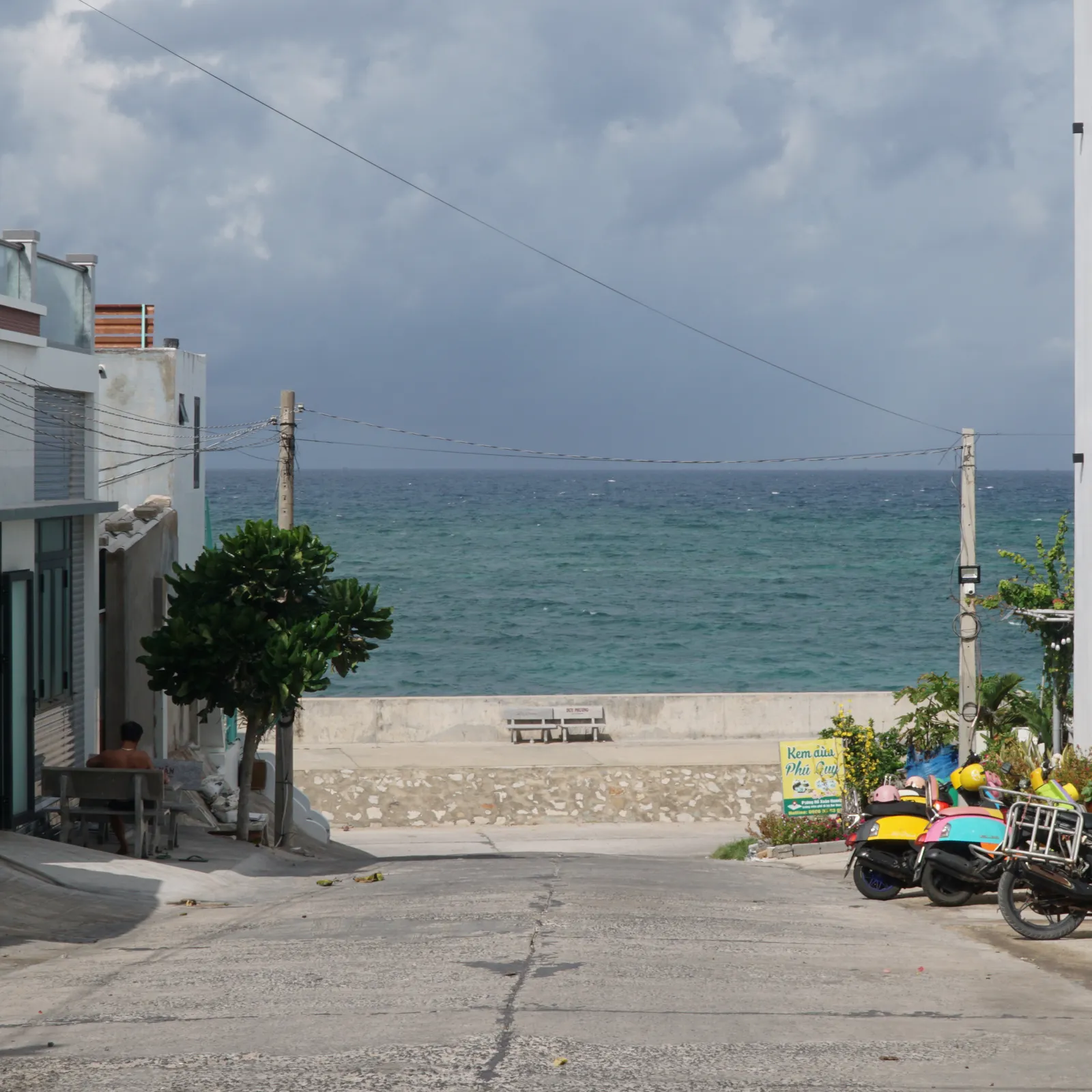 A street in Phu Quy running down to the sea wall, scooters parked by a coconut ice cream sign, the sea beyond