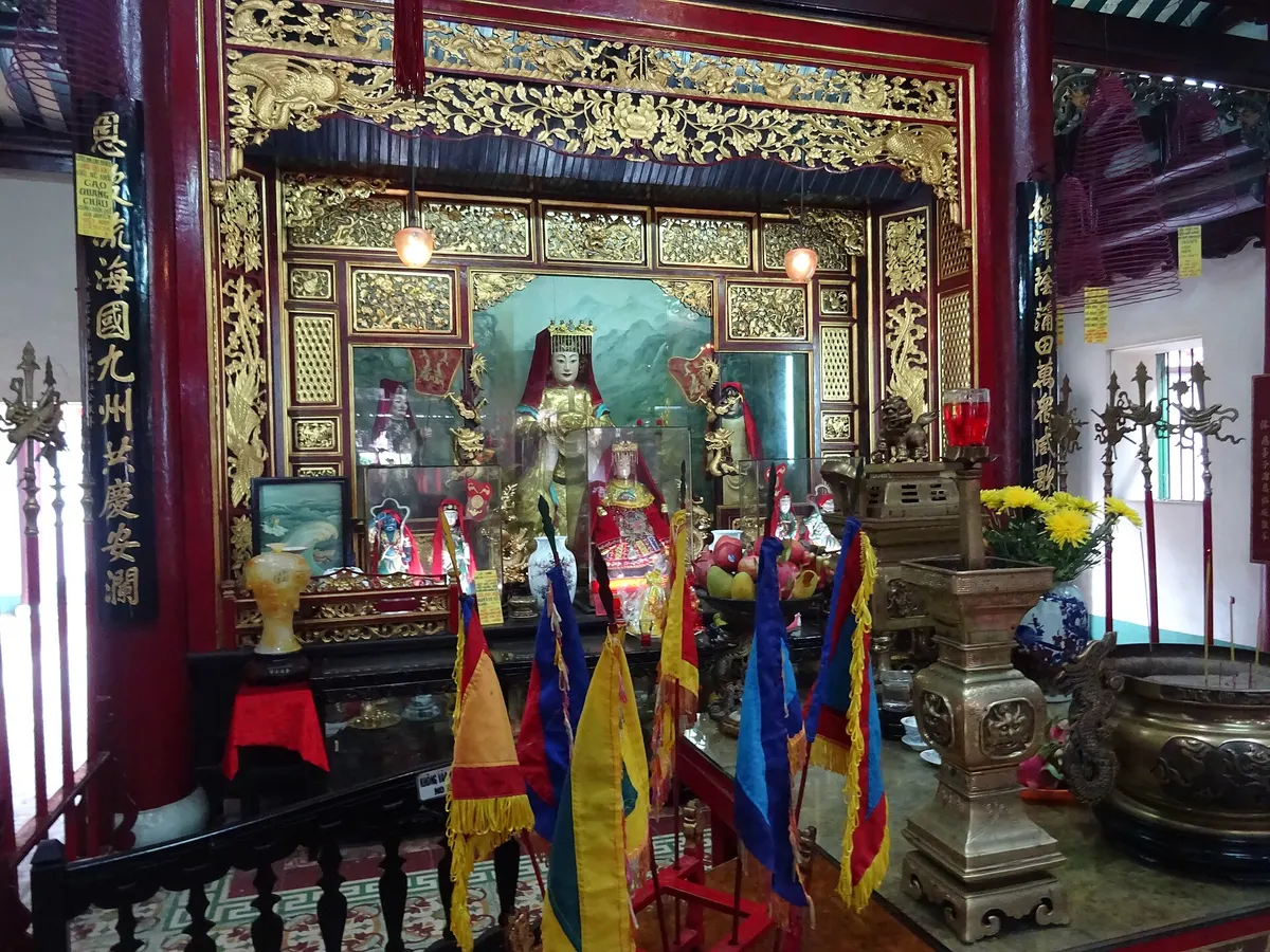 A gilded altar in Phuc Kien Assembly Hall with a statue of a goddess, colourful flags and offerings in front