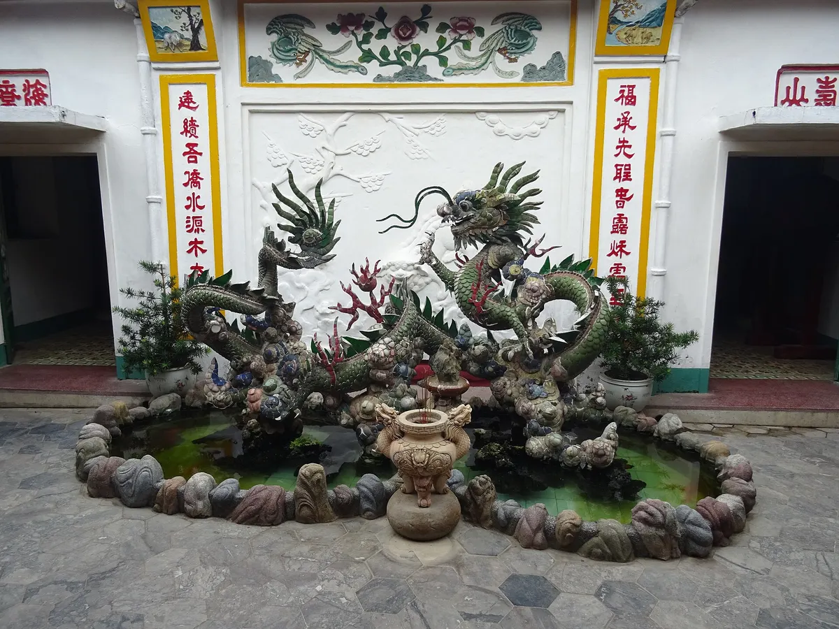 A dragon sculpture fountain in a small pond in front of a white wall with red and yellow calligraphy panels