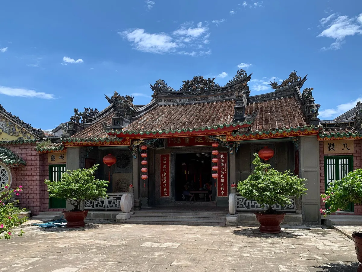 The main hall of Phuc Kien Assembly Hall with a curved tiled roof and red lanterns, potted bonsai trees in front and a paved courtyard