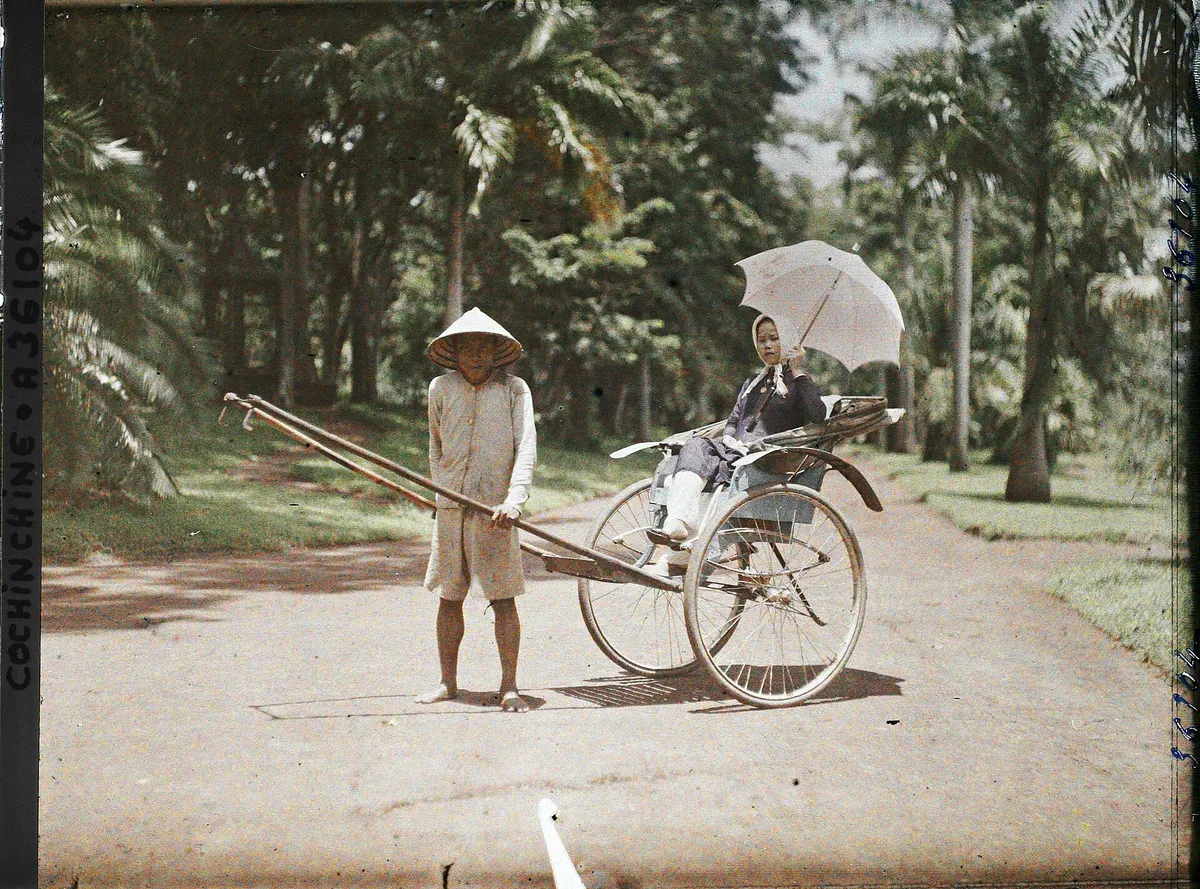 An early colour photograph of a woman with a parasol in a rickshaw on a path through the botanical garden, the puller standing beside it