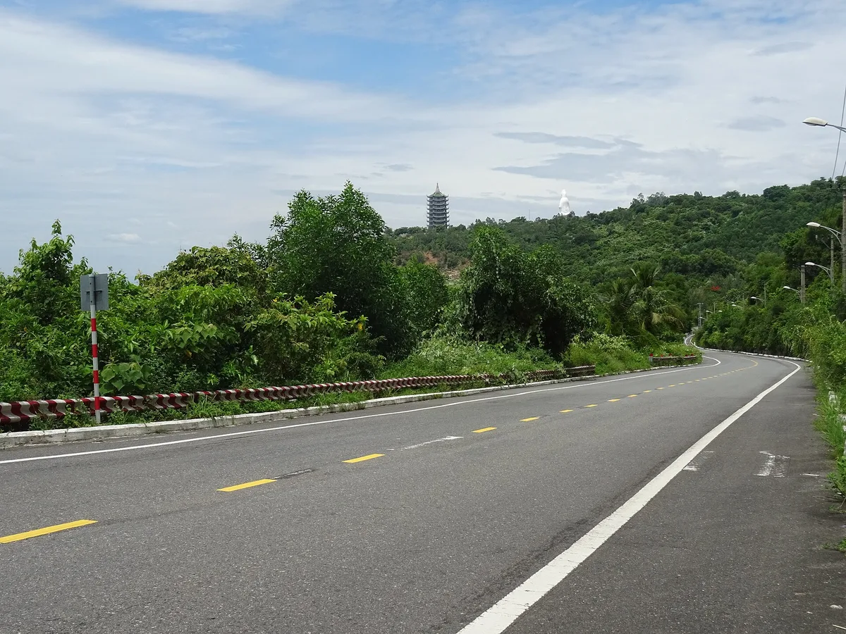 Hoang Sa road on Son Tra curving through green hills, with the pagoda tower and white Lady Buddha of Linh Ung in the distance