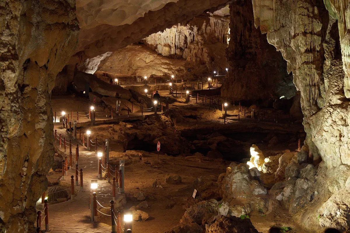 A paved walkway lined with lamp posts and rope barriers winds across the floor of a wide cave chamber