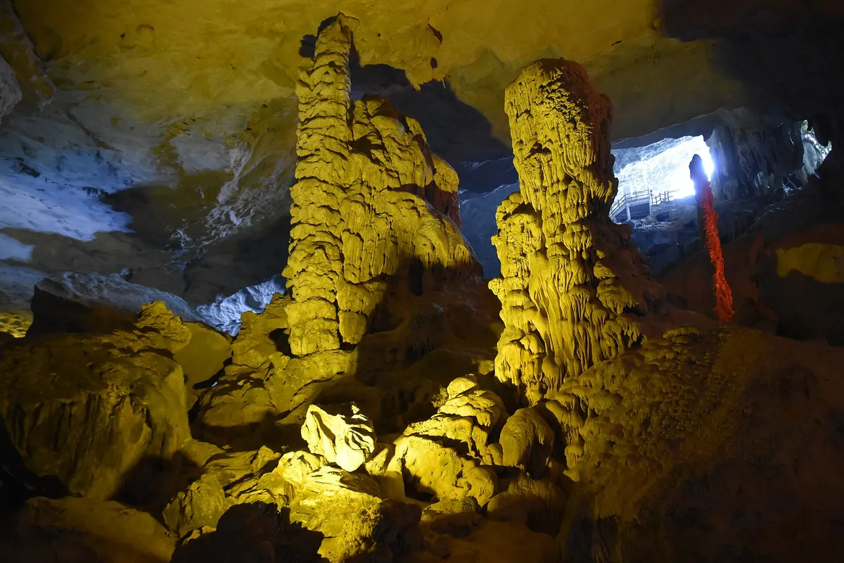 Two tall yellow-lit stalagmite columns rise from the cave floor, with daylight from an opening behind them