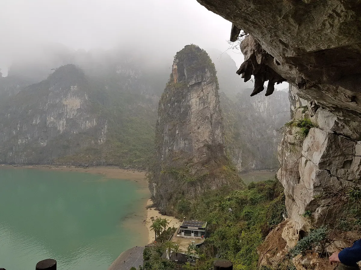 View from the mouth of Sung Sot Cave over a green lagoon and a tall limestone pillar half lost in mist