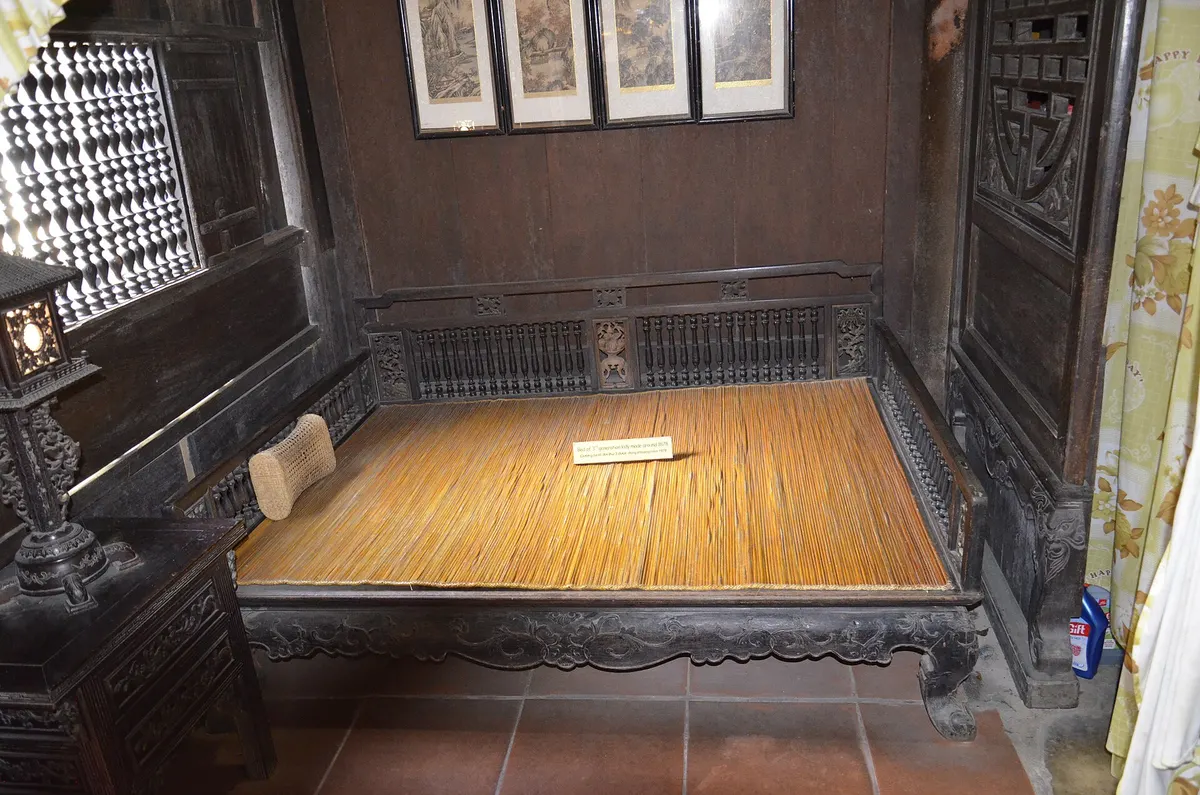 A low wooden daybed with a woven mat in a dark panelled room, framed pictures above it and a lattice window to one side