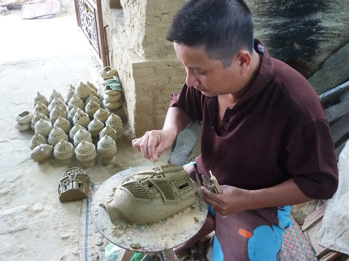 A potter carving fine detail into a clay model ship on a wheel, with rows of small perforated clay lamps on the floor beside him