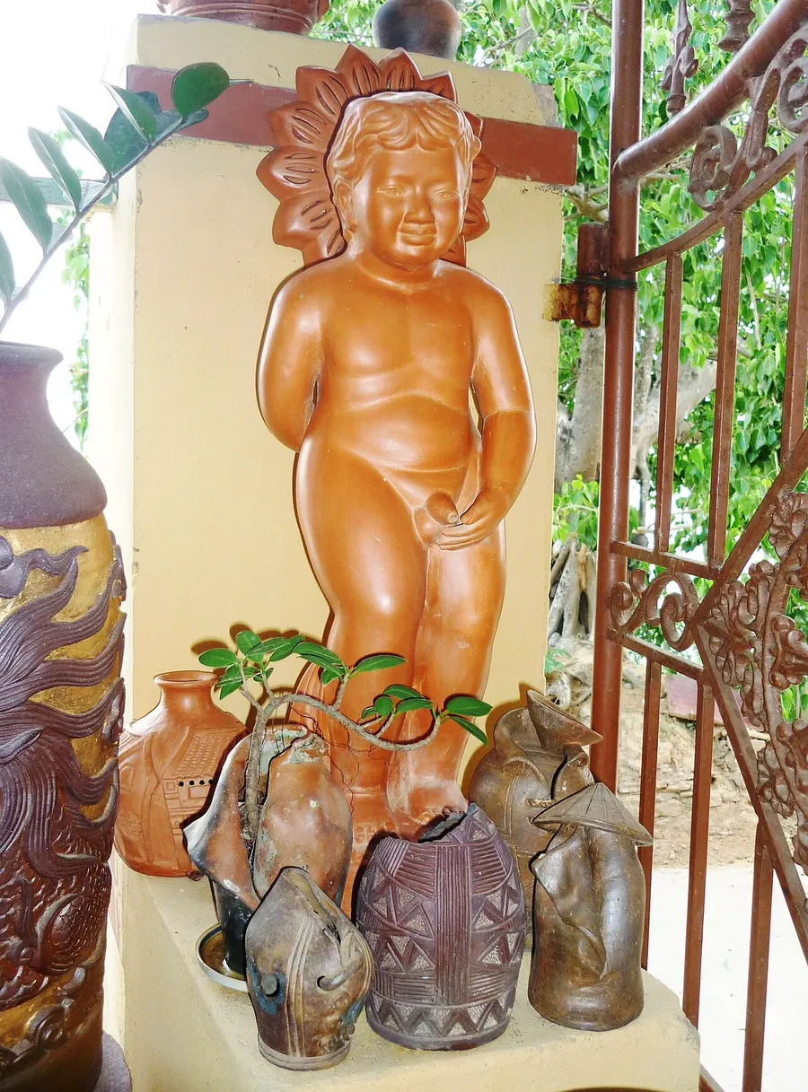 A smiling terracotta statue of a small boy beside a garden gate, with dark-glazed pots at its feet