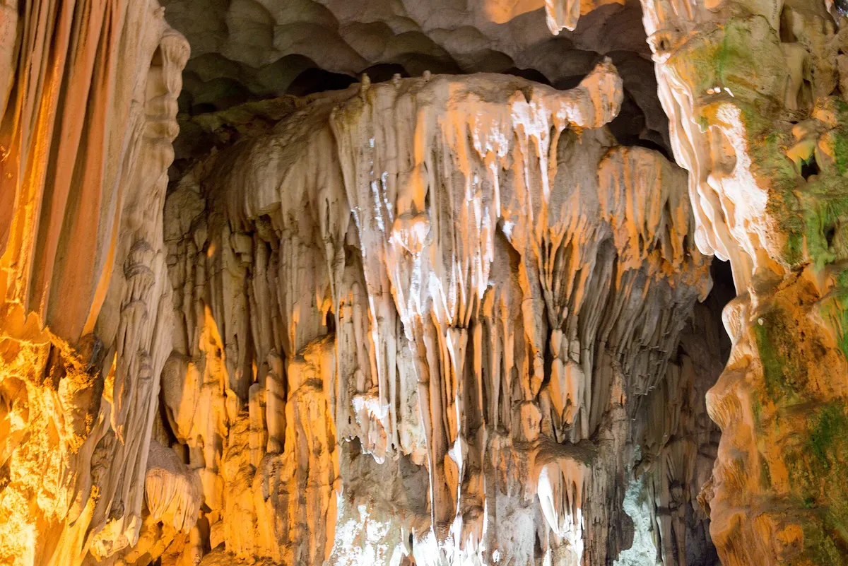 Pale stalactites hang in folds from a cave ceiling, lit orange from below