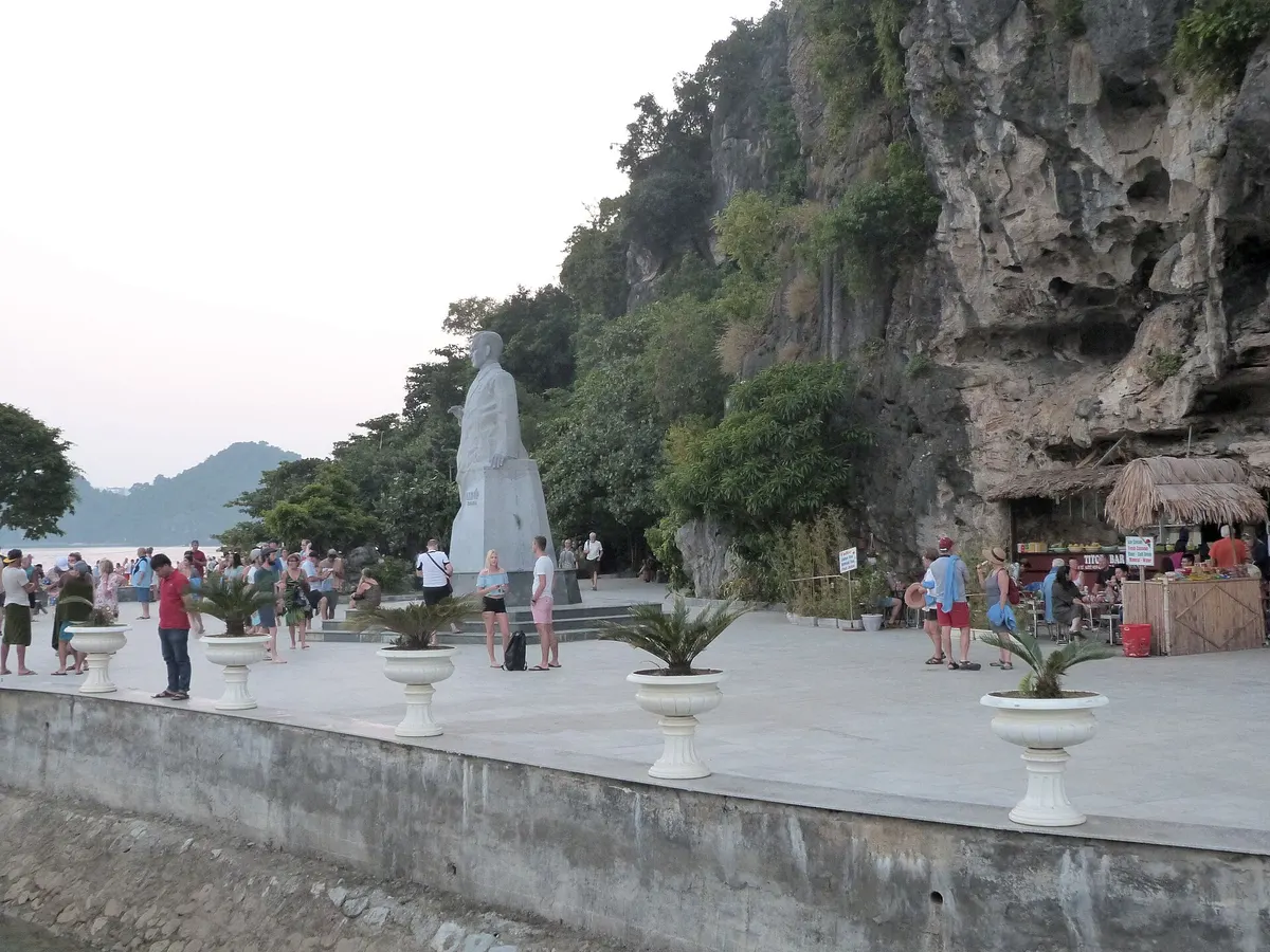 A white statue on a paved waterfront below the cliffs of Ti Top Island, with visitors and food stalls nearby