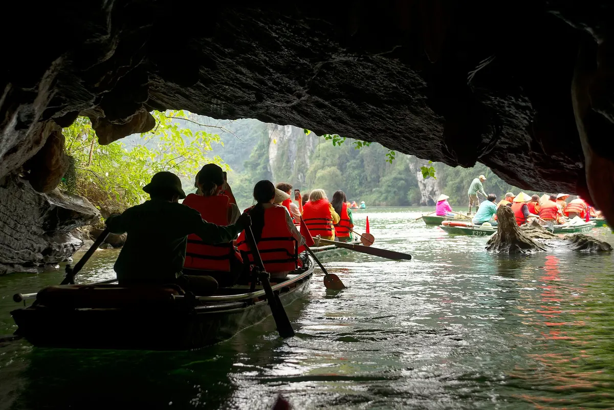 Visitors in orange life jackets rowing out of a low cave mouth into daylight, with more boats ahead