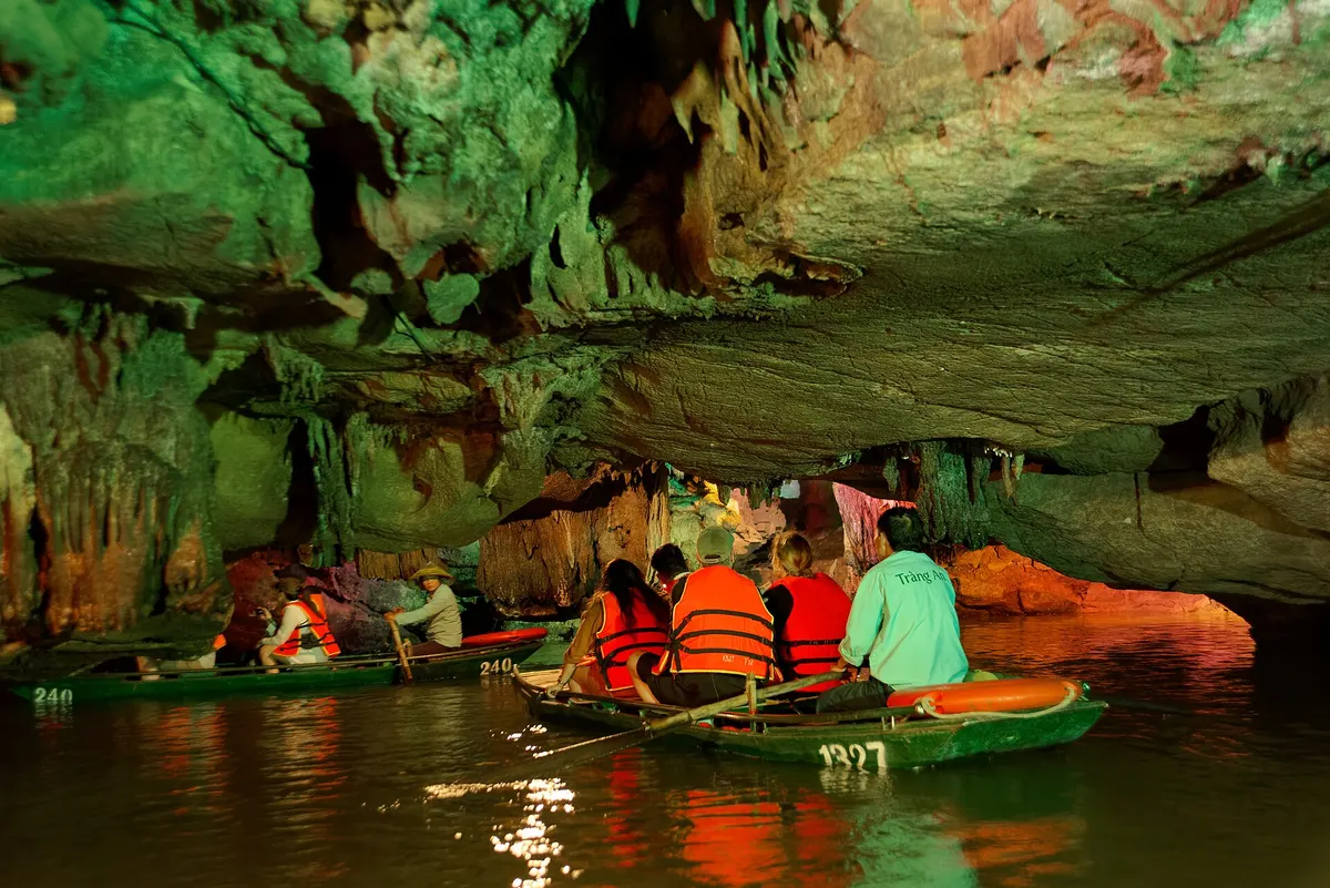 Rowing boats with visitors in orange life jackets passing under hanging stalactites in a lit cave