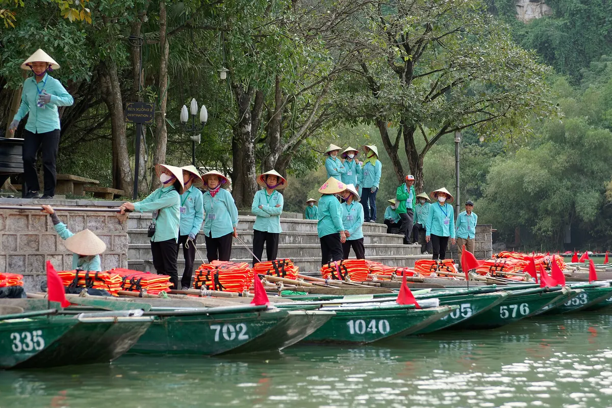 Boat rowers in teal uniforms and conical hats standing beside numbered rowing boats at the Trang An dock