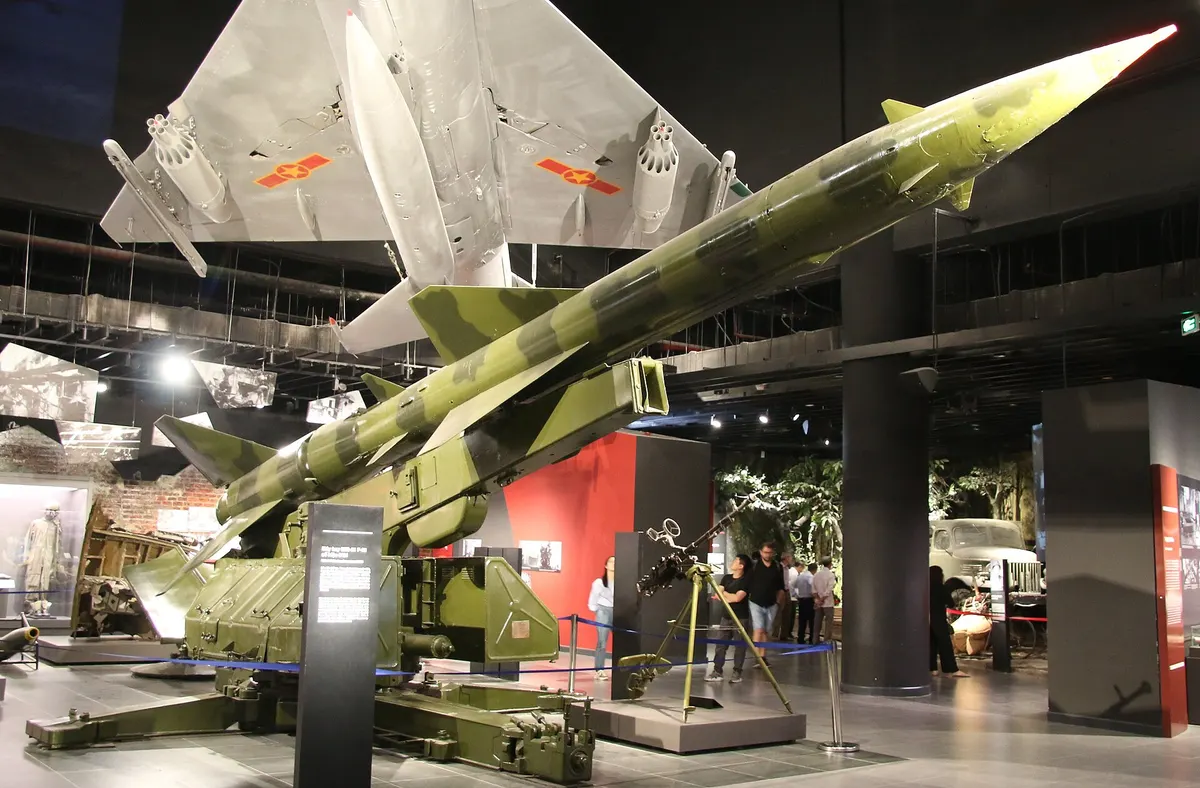 A camouflage-green SA-2 missile on its launcher indoors, with a silver jet fighter hanging above
