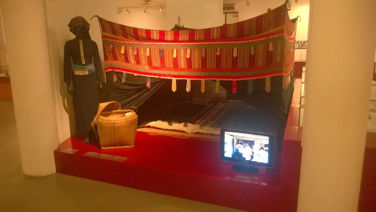 A display of a woman's dark costume, a basket and a woven cloth hung behind, with a small screen playing video