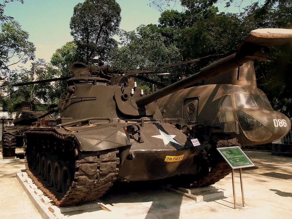 An olive-green tank and a twin-rotor helicopter displayed under trees in the museum grounds