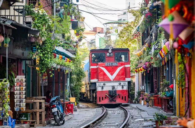 A red diesel train rolling down the narrow Hanoi Train Street between colorful houses